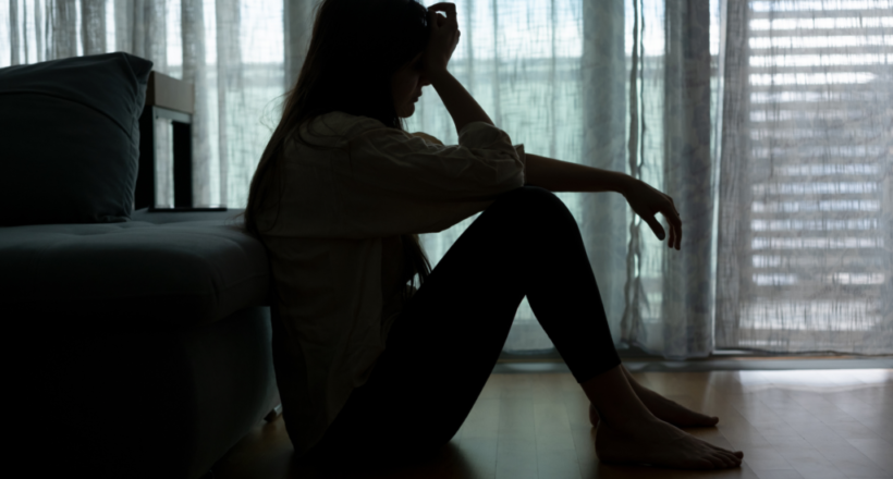 Silhouette of a woman sitting on the floor, holding her head in a dimly lit room, representing anxiety or depression.