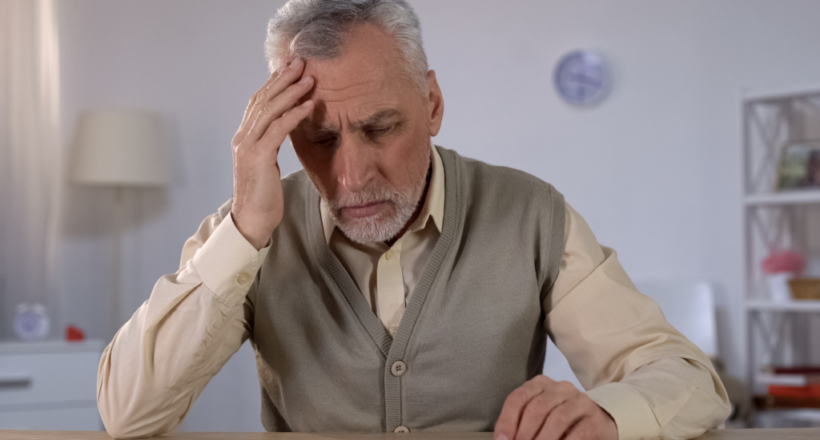 Elderly man showing signs of cognitive stress, resting head in hand while looking at scattered papers.