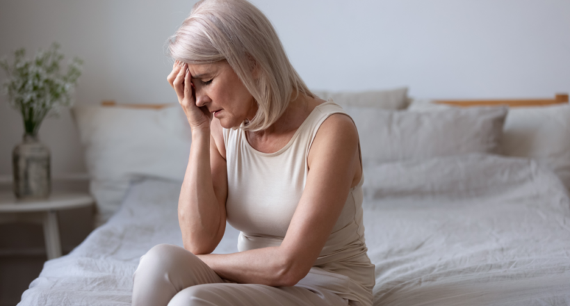 Woman experiencing menopausal symptoms sits on bed holding head.