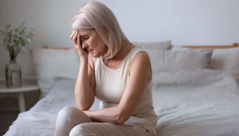 Woman experiencing menopausal symptoms sits on bed holding head.