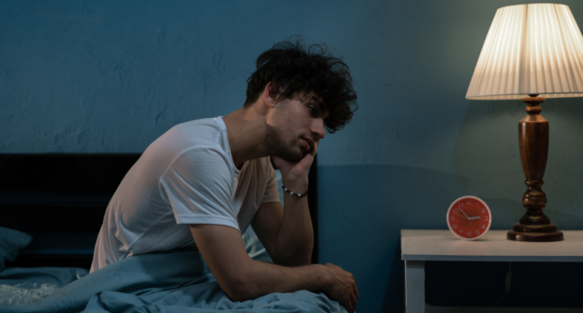 Exhausted young man experiencing disrupted sleep sits on bed at night.