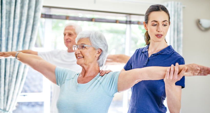 Physical therapist guides elderly woman and man doing arm exercises in a bright room.