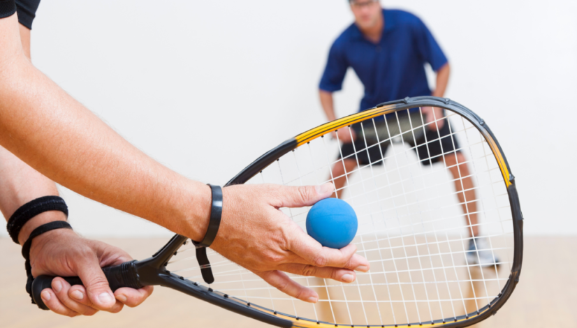 Close-up of a racquetball serve with a blue ball and racquet, opponent in the background ready to play on an indoor court.