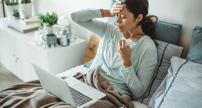 how-to-prevent-common-cold Sick woman in bed with tissue and laptop, experiencing common cold symptoms.