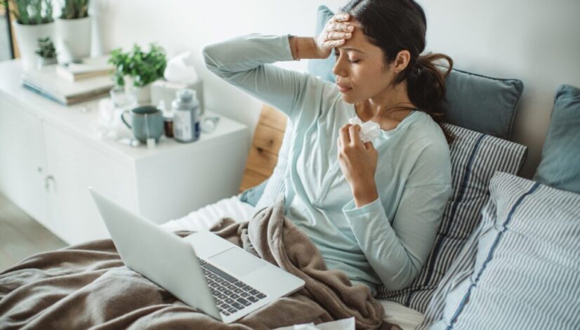 Sick woman in bed with tissue and laptop, experiencing common cold symptoms.