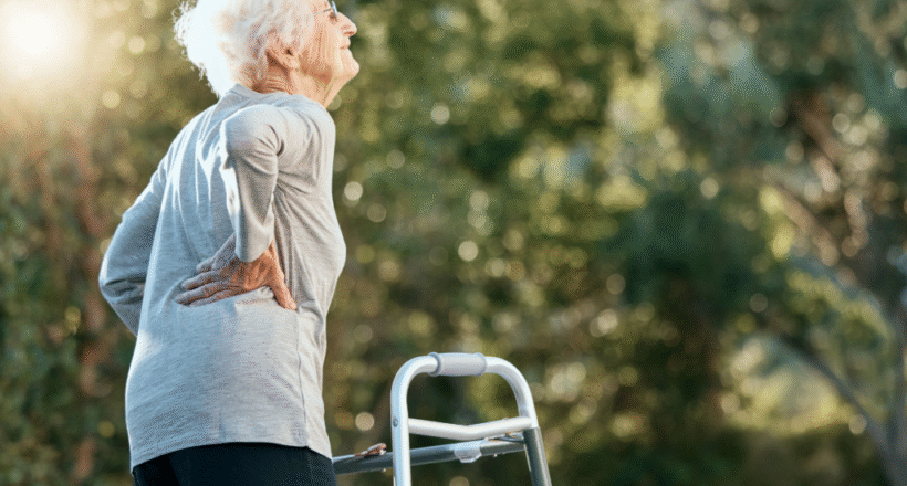 Senior woman outdoors holding her lower back in discomfort beside a walker.