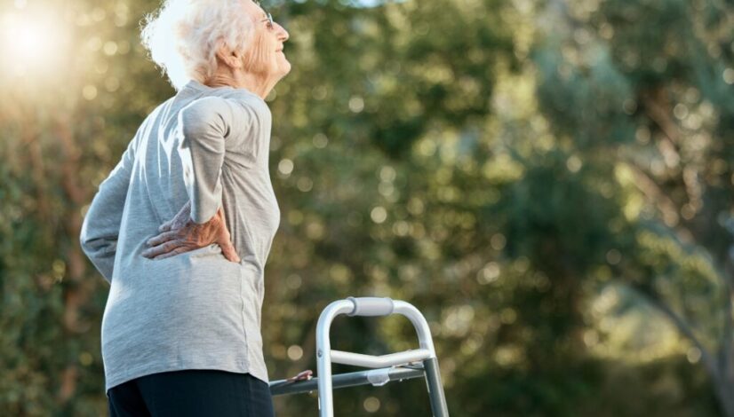 Senior woman outdoors holding her lower back in discomfort beside a walker.
