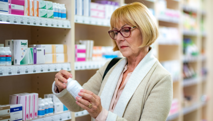 Senior woman reading over-the-counter medicine label in pharmacy aisle, checking ingredients before purchase.