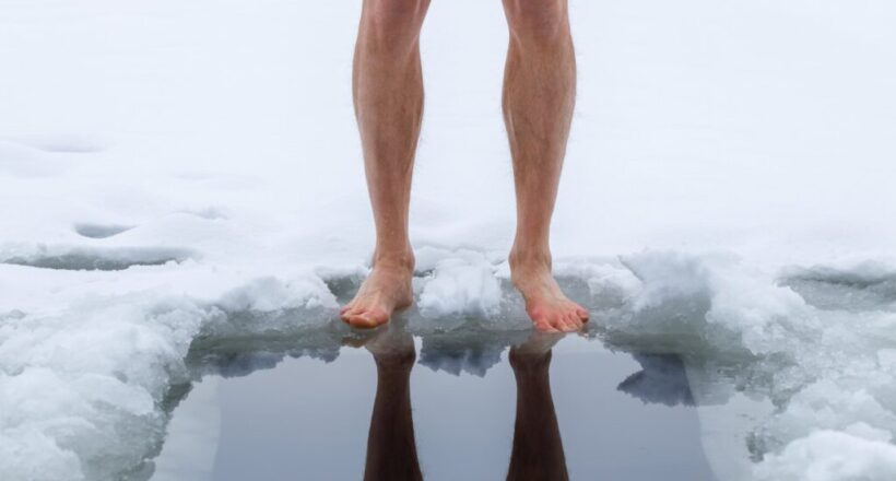 Barefoot person standing at the edge of an icy plunge hole in the snow.
