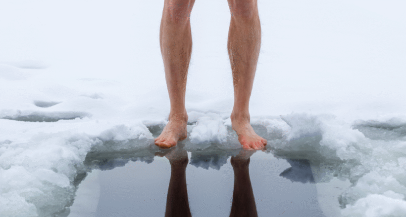 cold-plunge-benefits Barefoot person standing at the edge of an icy plunge hole in the snow.