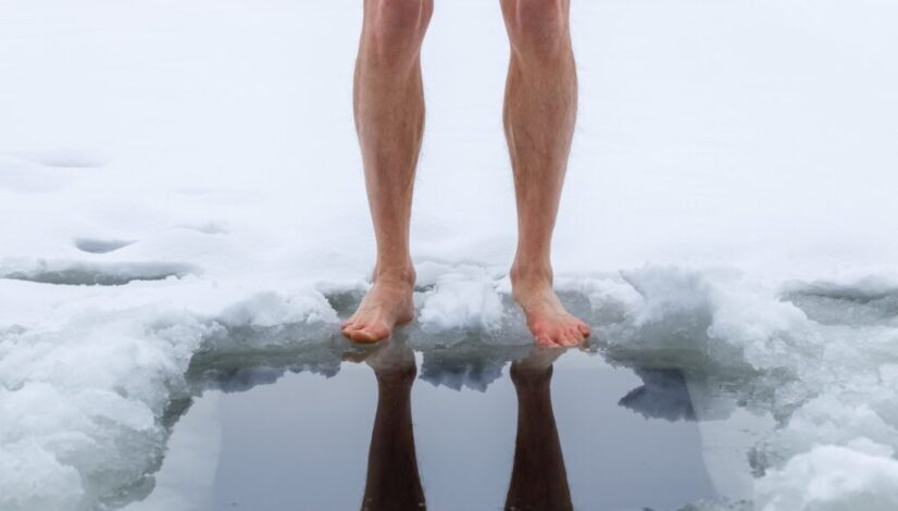 Barefoot person standing at the edge of an icy plunge hole in the snow.