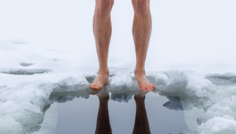Barefoot person standing at the edge of an icy plunge hole in the snow.