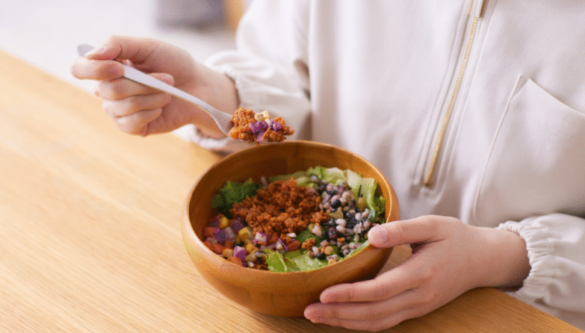 Person eating a healthy salad bowl with beans, lettuce, and plant-based protein at a wooden table.