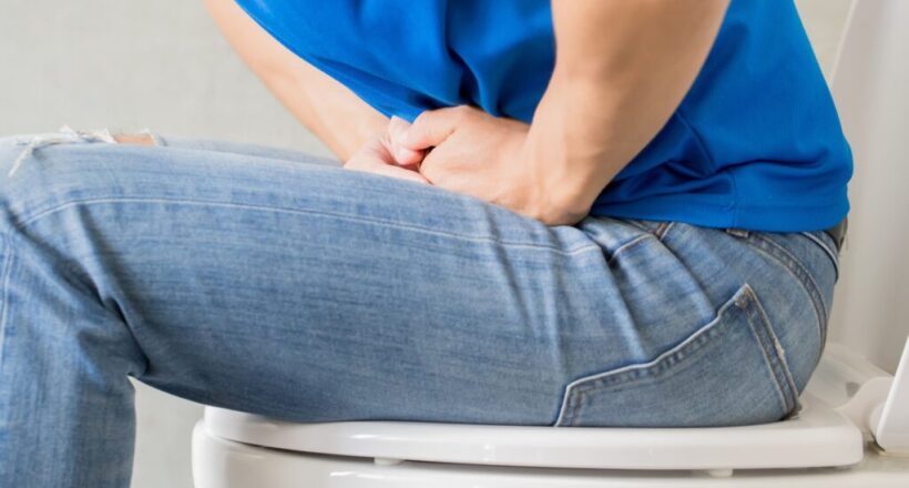 Person in jeans with and blue shirt sits on toilet.