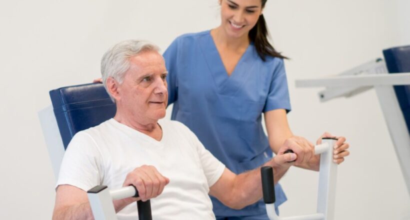 Physical therapist smiles and guides elderly male patient using exercise equipment.